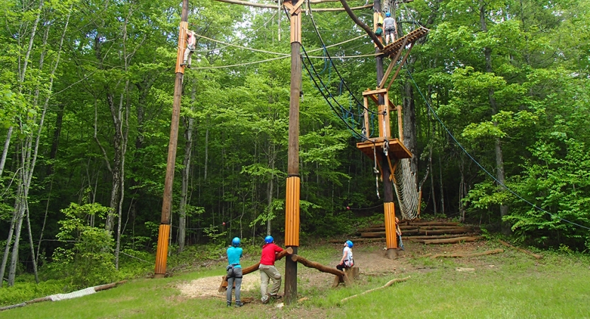 A group of people look up at another person who is navigating a high ropes course. Everyone is wearing safety gear. 